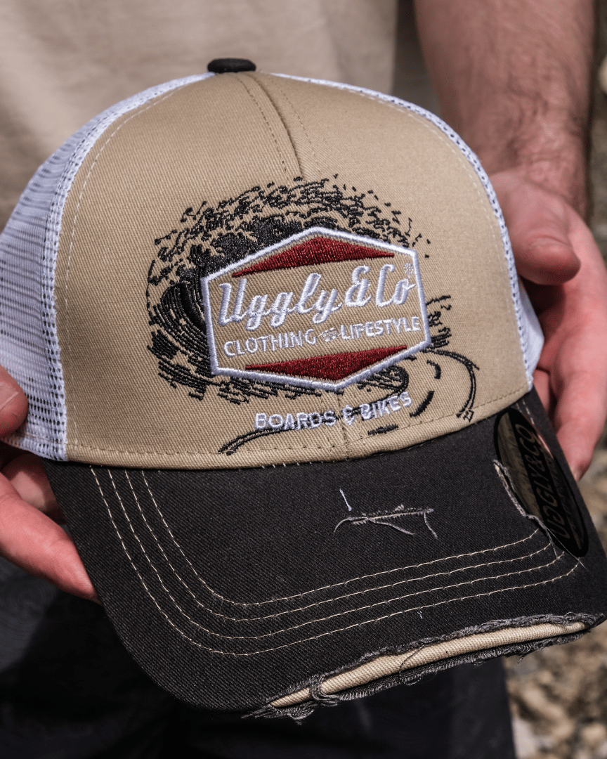 A model holding a beige and brown trucker cap with a diamond Uggly&Co logo and wave texture design, featuring a white mesh back and a curved peak.