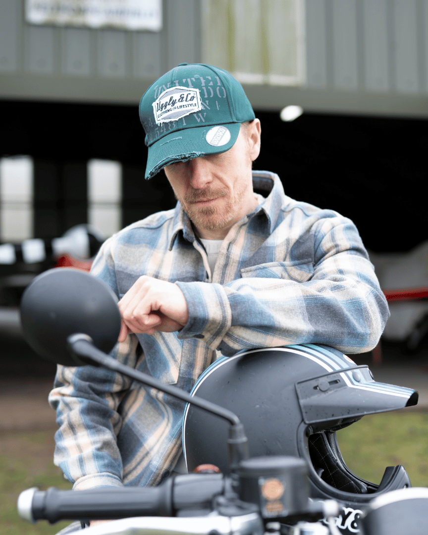 Man looking down while wearing a green 'Aviation' Diamond Trucker Hat with distressed black and white Uggly&Co logo and plaid jacket sitting on a motorcycle on an airfield