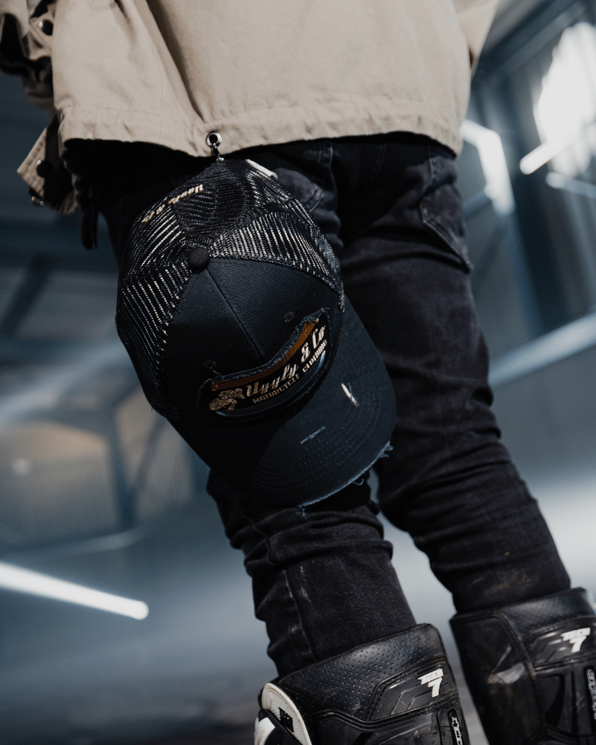 An all-black trucker hat with a vintage-inspired flat track racing logo in yellow and blue on the front hanging from a model's pocket with a blurred background