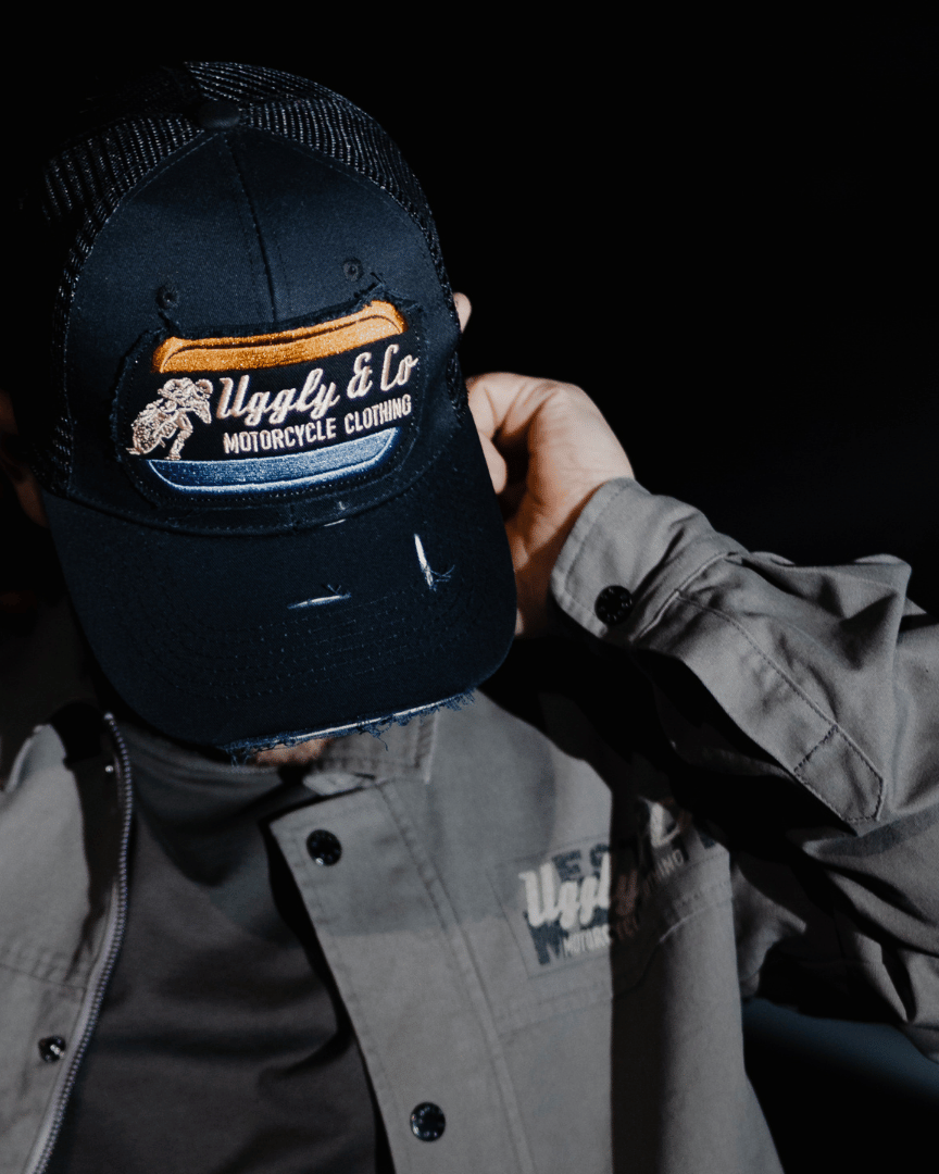 A model looking down while wearing an all-black trucker hat with a vintage-inspired flat track racing logo in yellow and blue on the front with a black background