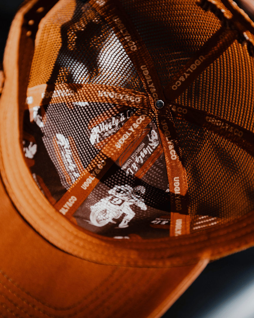Close up of the inside of a brown trucker hat with an orange brim, featuring Uggly&Co branding on the seams at night