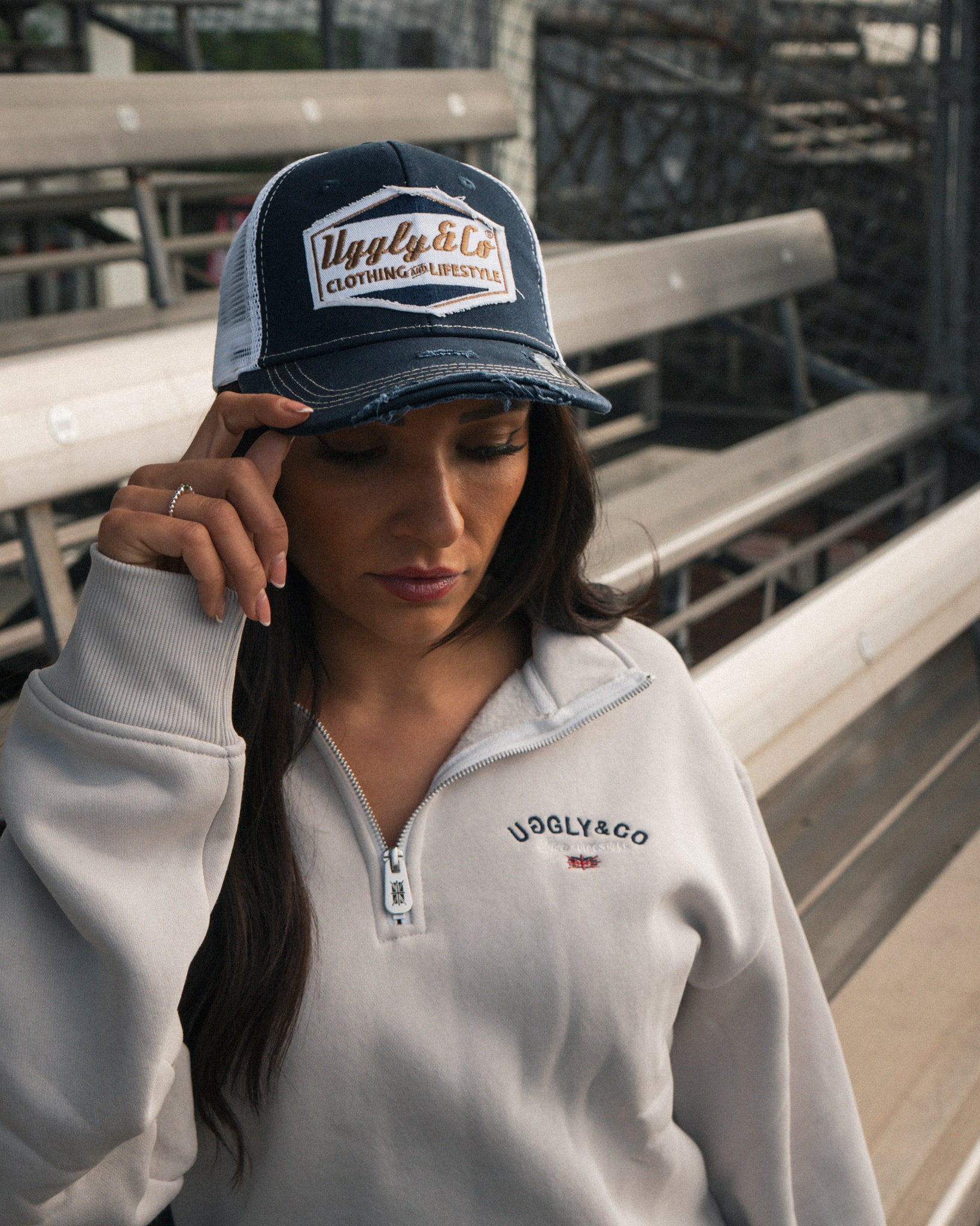 Female model wearing the Uggly&Co Night Blue and Gold Diamond Trucker Hat in a motorsport grandstand