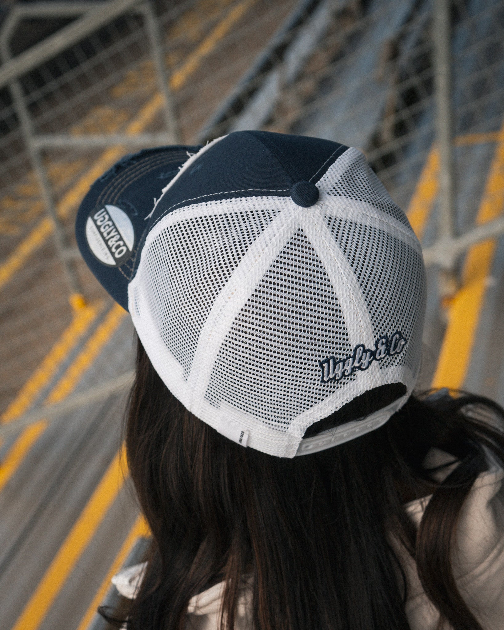 Uggly&Co Night Blue and Gold Diamond Trucker Hat worn by a female model in a racing grandstand