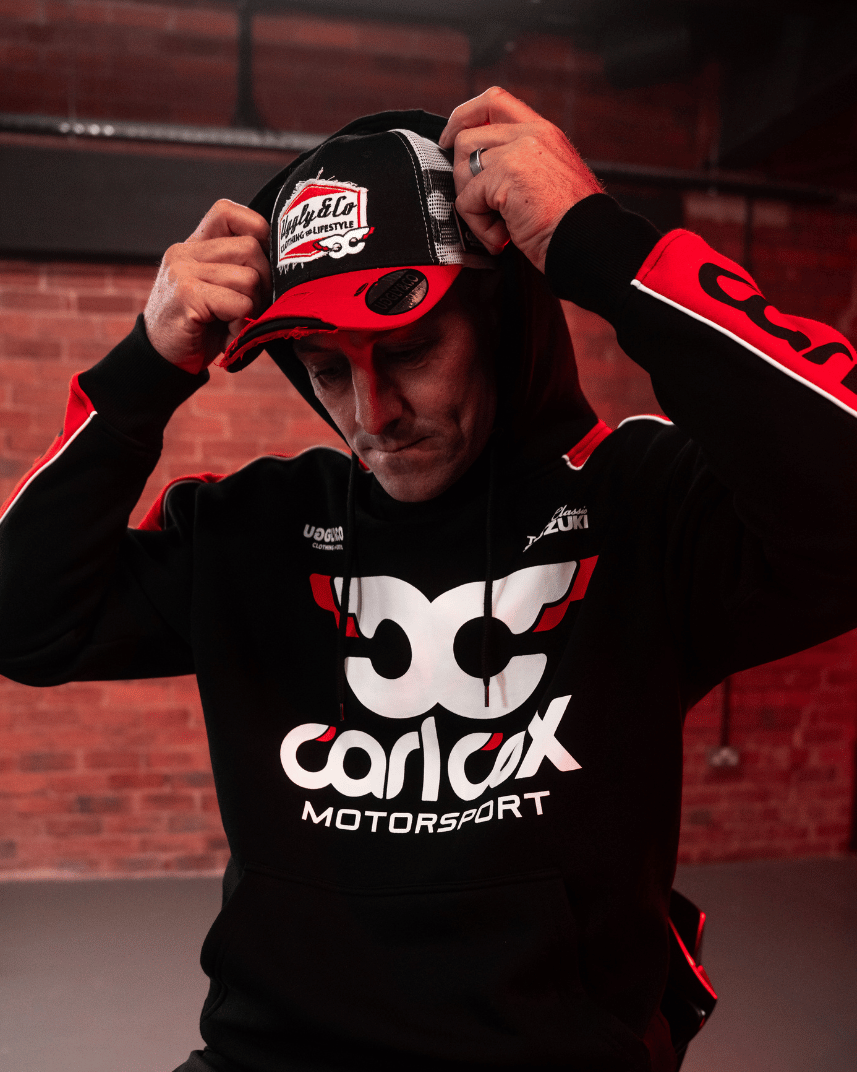 Josh Brookes putting his hood up while wearing a black hoodie with red sleeves and trucker hat featuring Carl Cox Motorsport branding in the north west 200 paddock