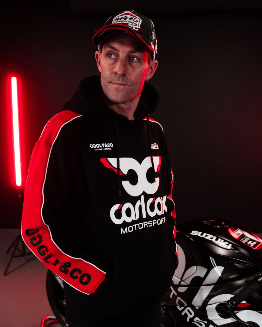 Josh Brookes wearing a black hoodie with red sleeves featuring Carl Cox Motorsport branding in front of his motorbike