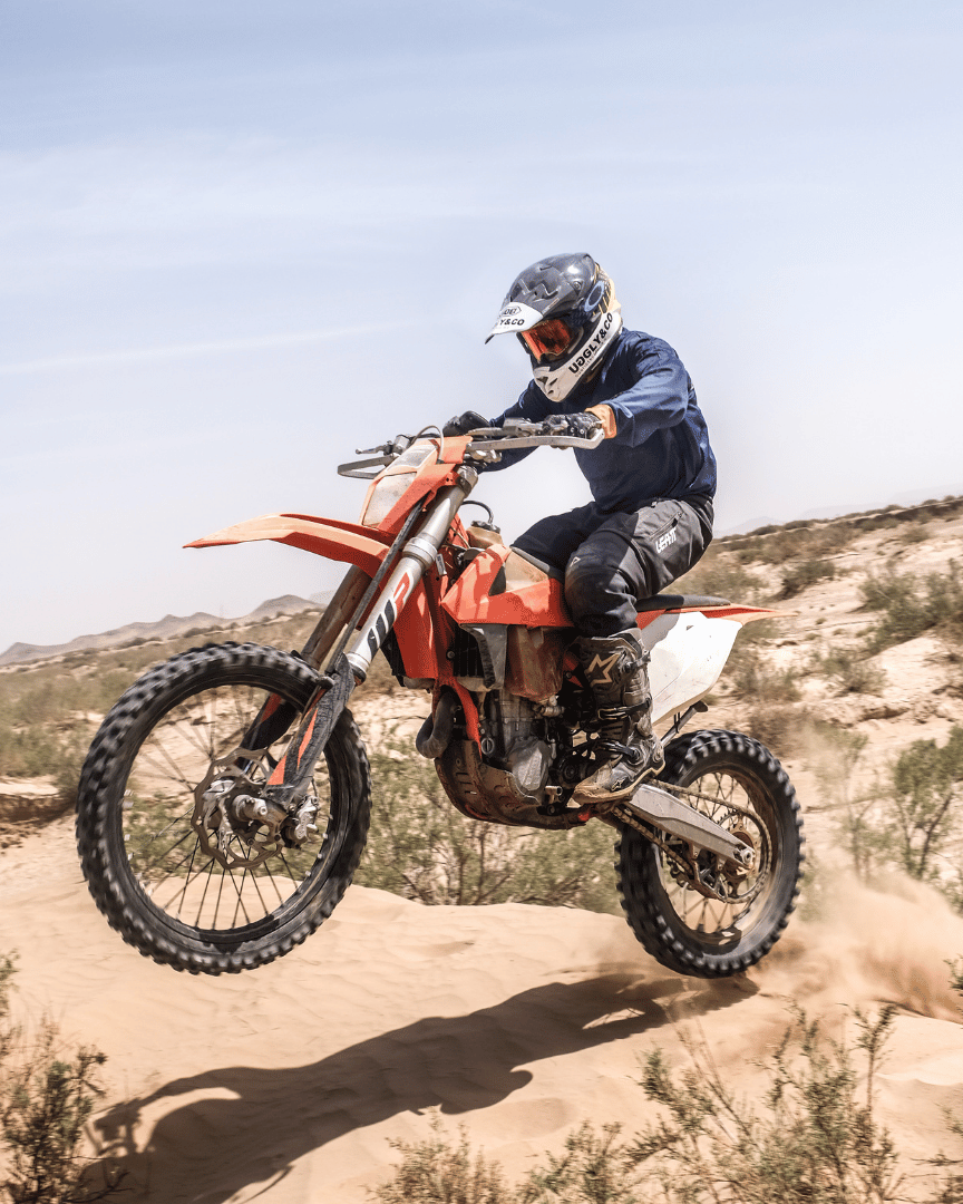 MX rider on a bike in the desert in a blue motocross jersey and the brand name 'Uggly&Co Motorcycle Clothing' on the front