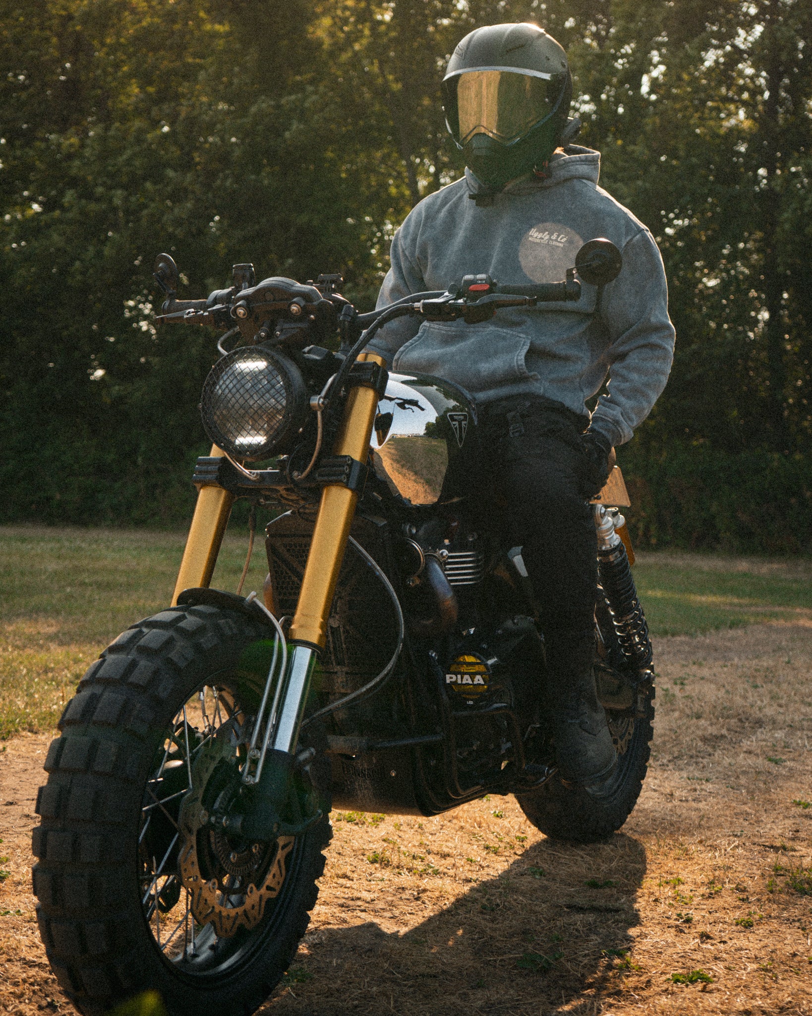 Golden hour lifestyle shot of a male rider in an acid wash hoodie on a Triumph Scrambler bike.