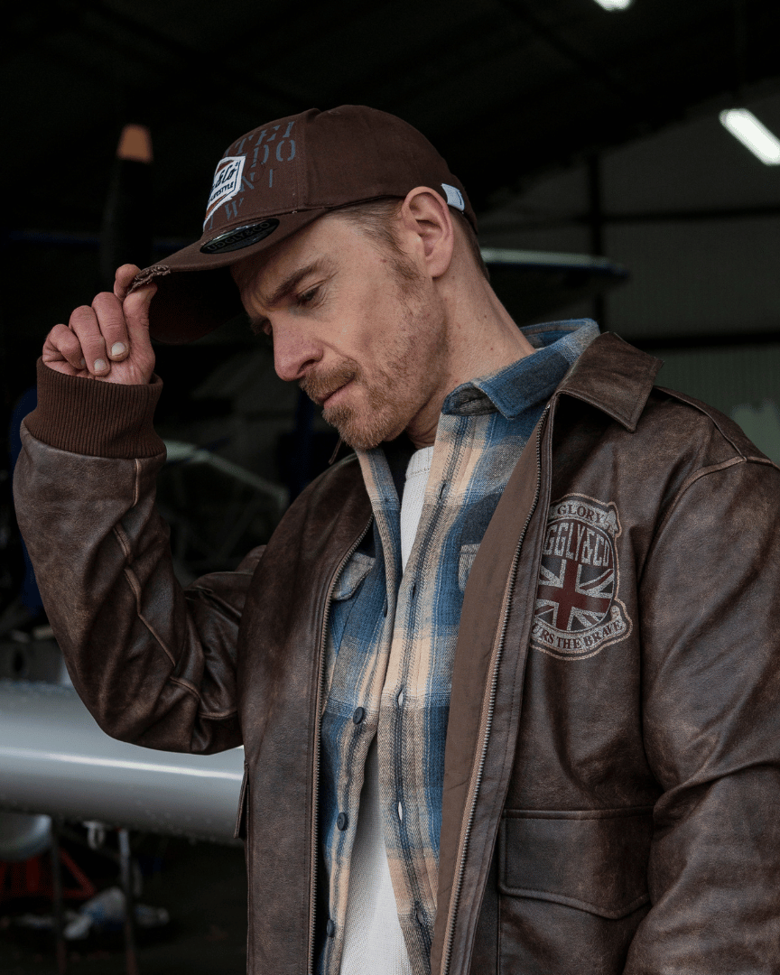 Man wearing a brown leather jacket and brown aviation trucker hat, standing in front of an airplane.