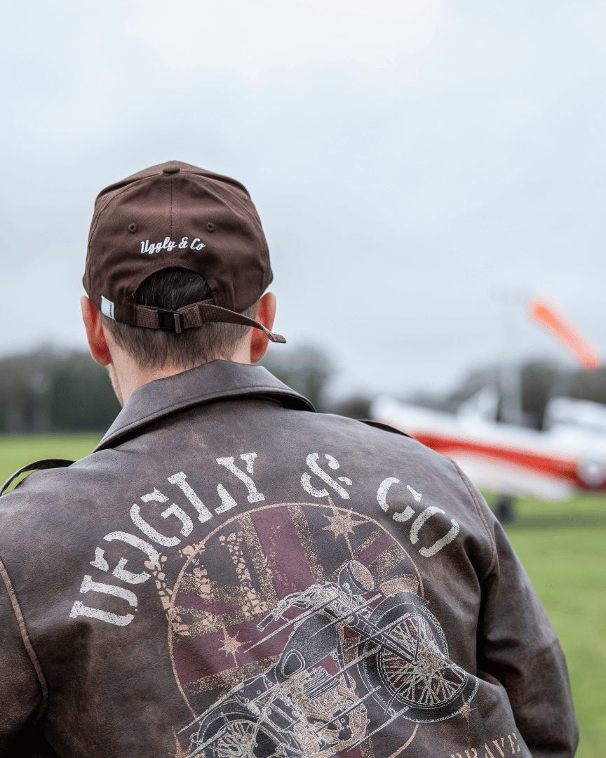 Rear view of a man wearing a brown jacket with 'Uggly & Co' branding, looking at an airplane on a grassy field.