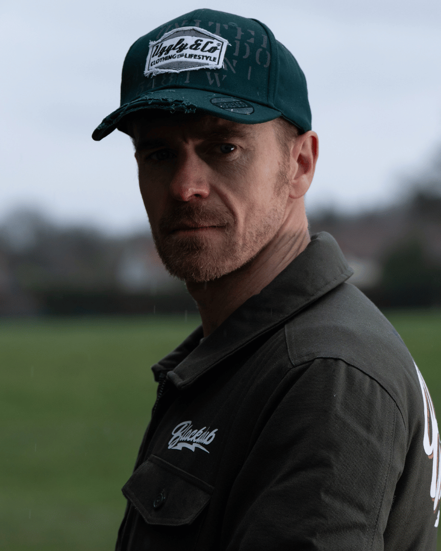 A man wearing a green 'Aviation' Diamond Trucker Hat with distressed black and white Uggly&Co logo on an airfield