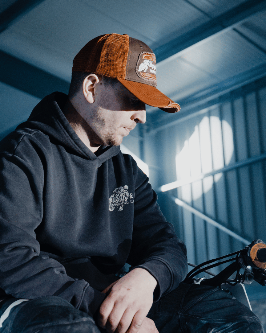 A model looking down to the right while wearing a brown trucker hat with an orange brim, featuring a flat track Uggly&Co logo patch on the front in a warehouse