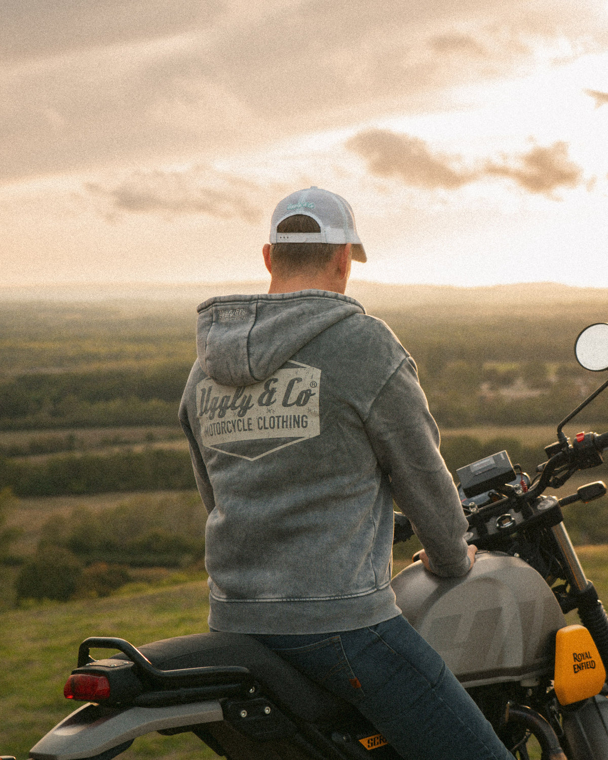 Lifestyle shot of a motorcycle rider in a retro Uggly&Co trucker hat with a distressed peak, beside a Royal Enfield Scram 411