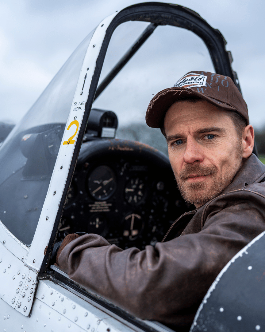 Man wearing a brown 'Aviation' Diamond Trucker Hat with the Uggly&Co logo on the front and brown leather jacket sitting in the cockpit of a small airplane.