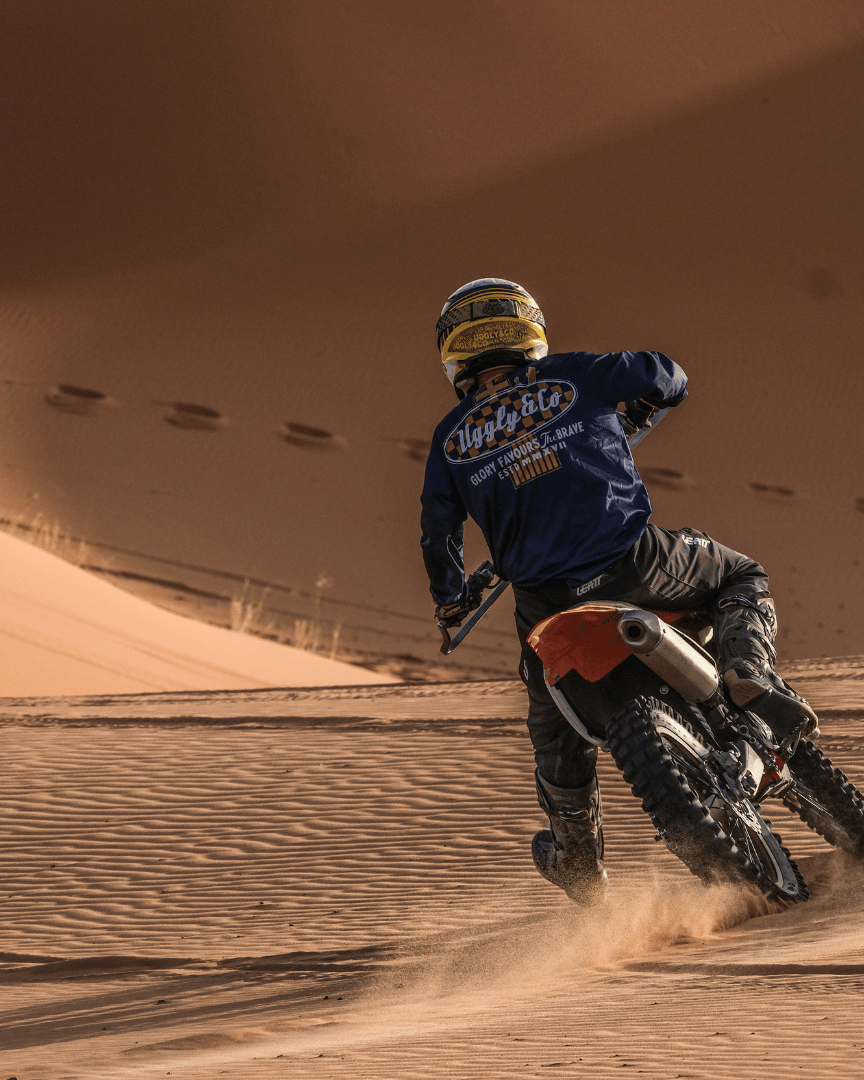 Riding a motocross bike in the desert in a blue motocross jersey and the brand name 'Uggly&Co Motorcycle Clothing' on the back