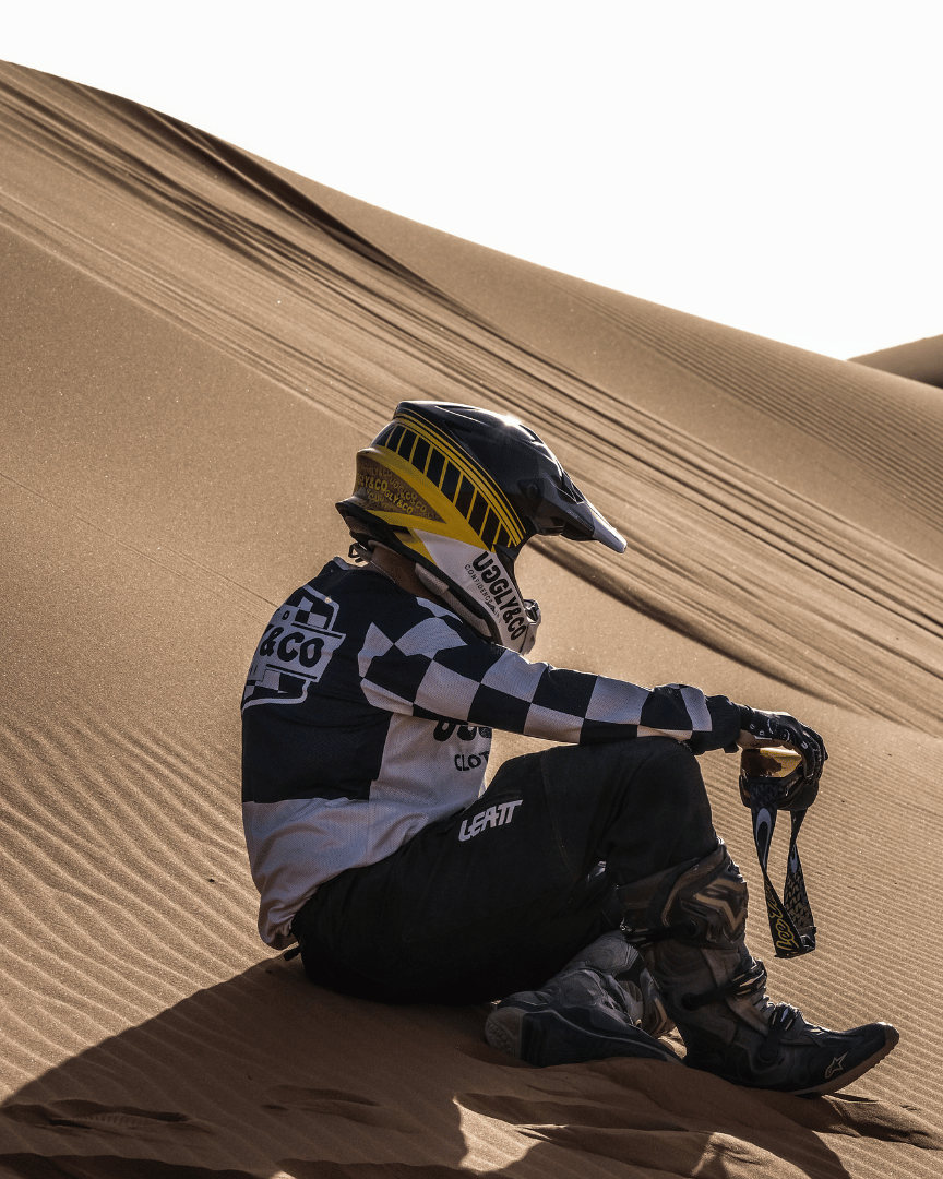 A motocross rider in full gear sitting on sand dunes in the desert wearing a chequered Uggly&Co MX shirt