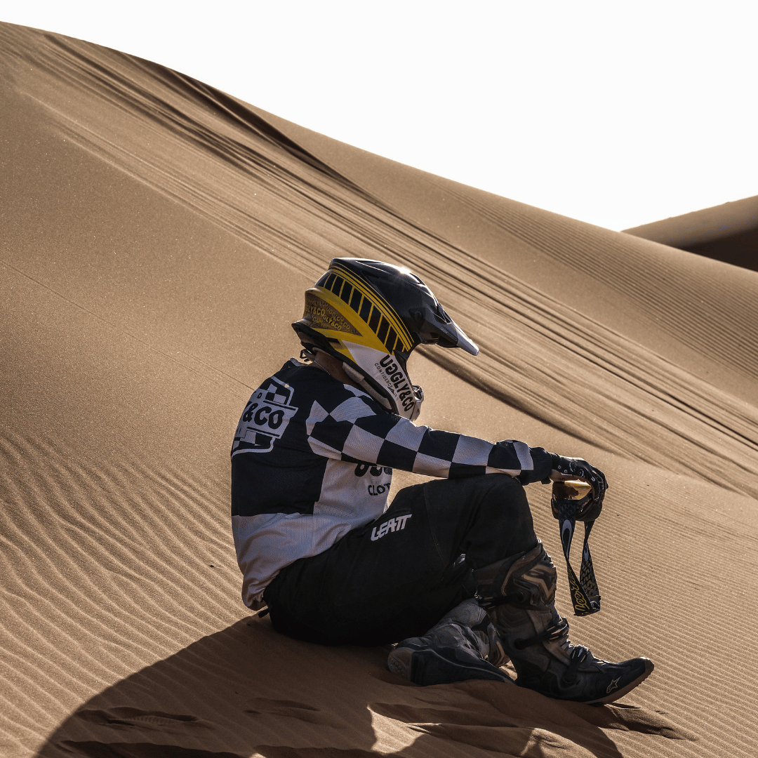 A motocross rider in full gear sitting on sand dunes in the desert wearing a chequered Uggly&Co MX shirt