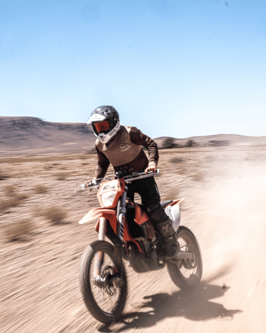 A person standing up while riding a motocross bike in the desert in an Uggly&Co MX shirt