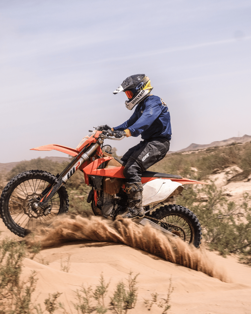 MX rider riding over sand dunes in the desert in a blue Uggly&Co motocross jersey