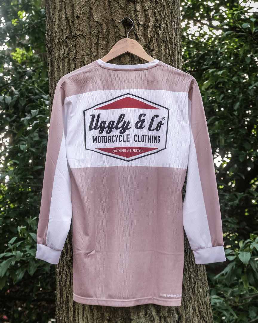 A beige motocross jersey with a diamond logo and the brand name 'Uggly&Co Motorcycle Clothing' on the front and back, hanging on a tree.
