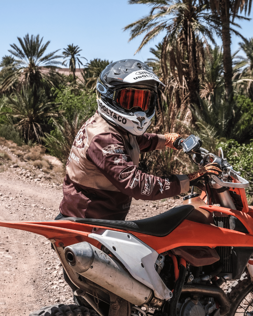 A person standing with a motocross bike in a brown Uggly&Co long-sleeve MX shirt in the desert
