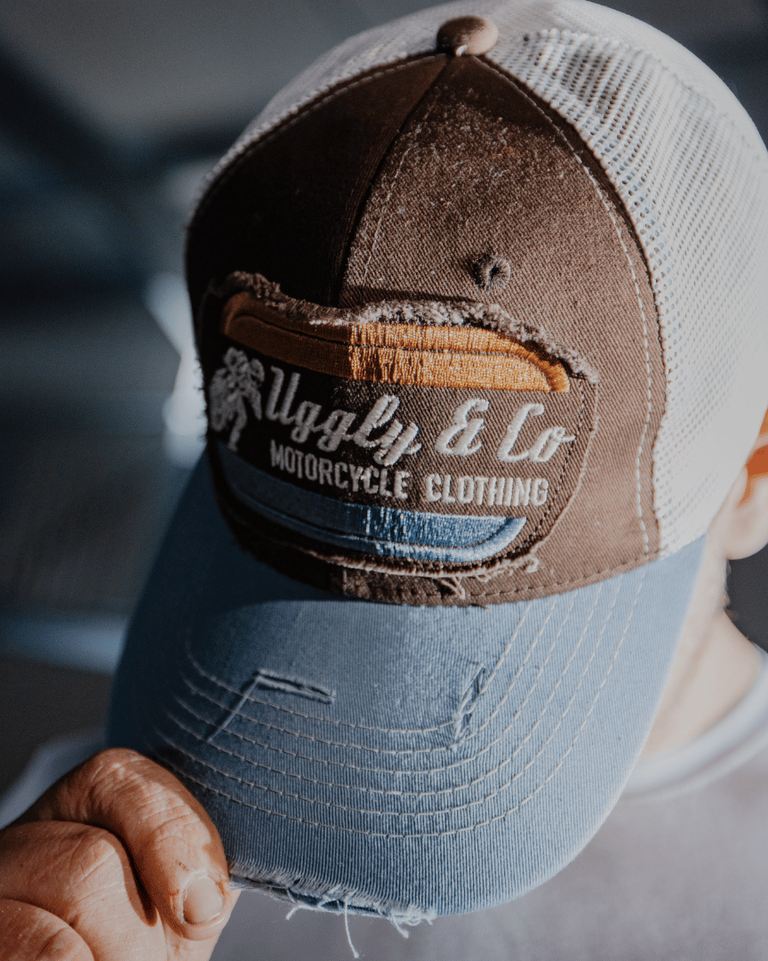 A model looking down while wearing a dusty blue trucker hat with a brown front panel, featuring a vintage flat track Uggly&Co logo