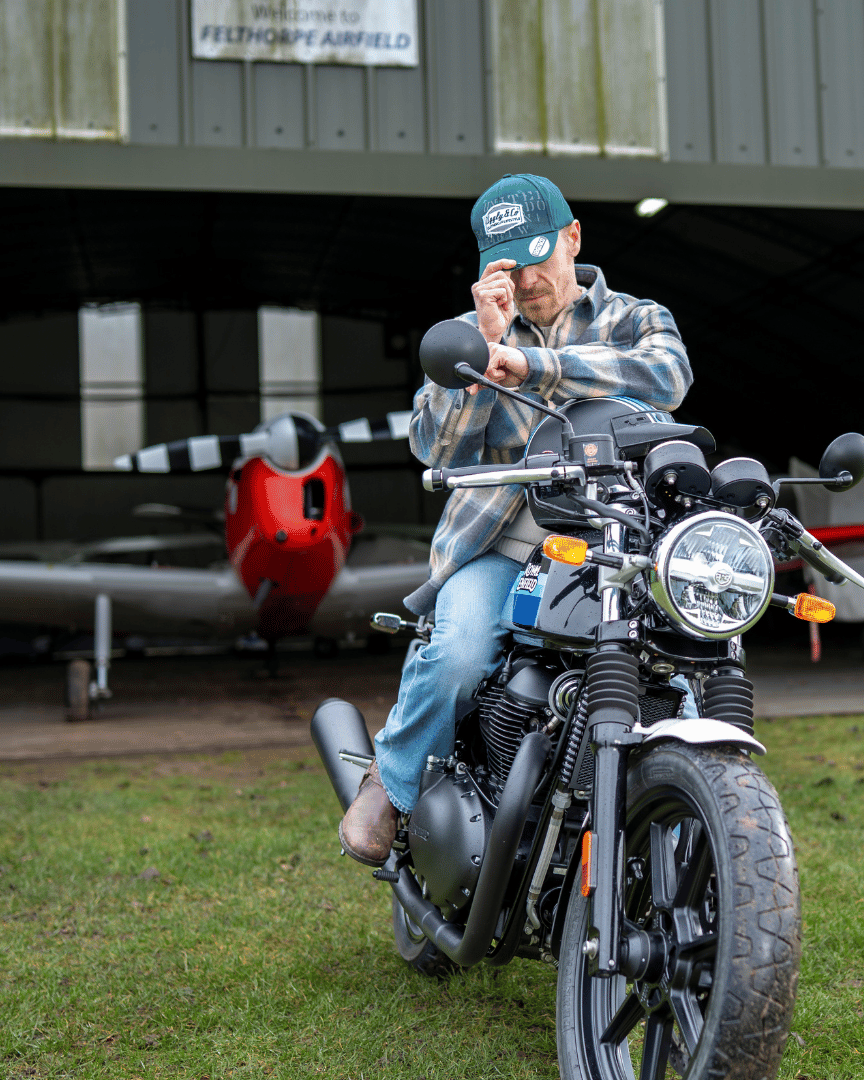 Man wearing a green 'Aviation' Diamond Trucker Hat with distressed black and white Uggly&Co logo and plaid jacket sitting on a motorcycle in front of a plane