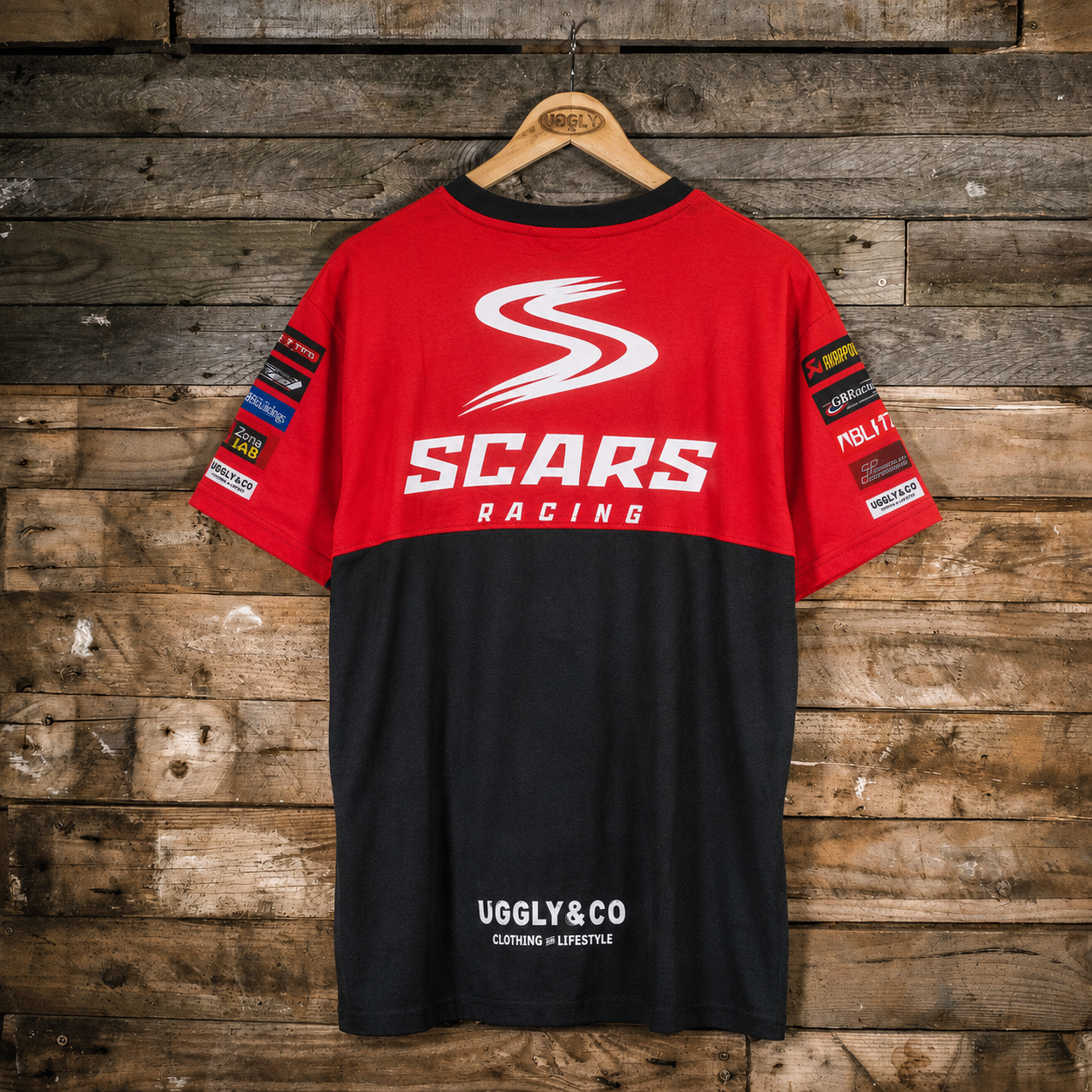A red and black short-sleeve t-shirt with 'SCARS RACING' printed on the back, hanging on a wooden hanger against a rustic wood plank wall. The shirt features sponsor patches on the sleeves and 'UGGLY&CO' printed on the lower back.