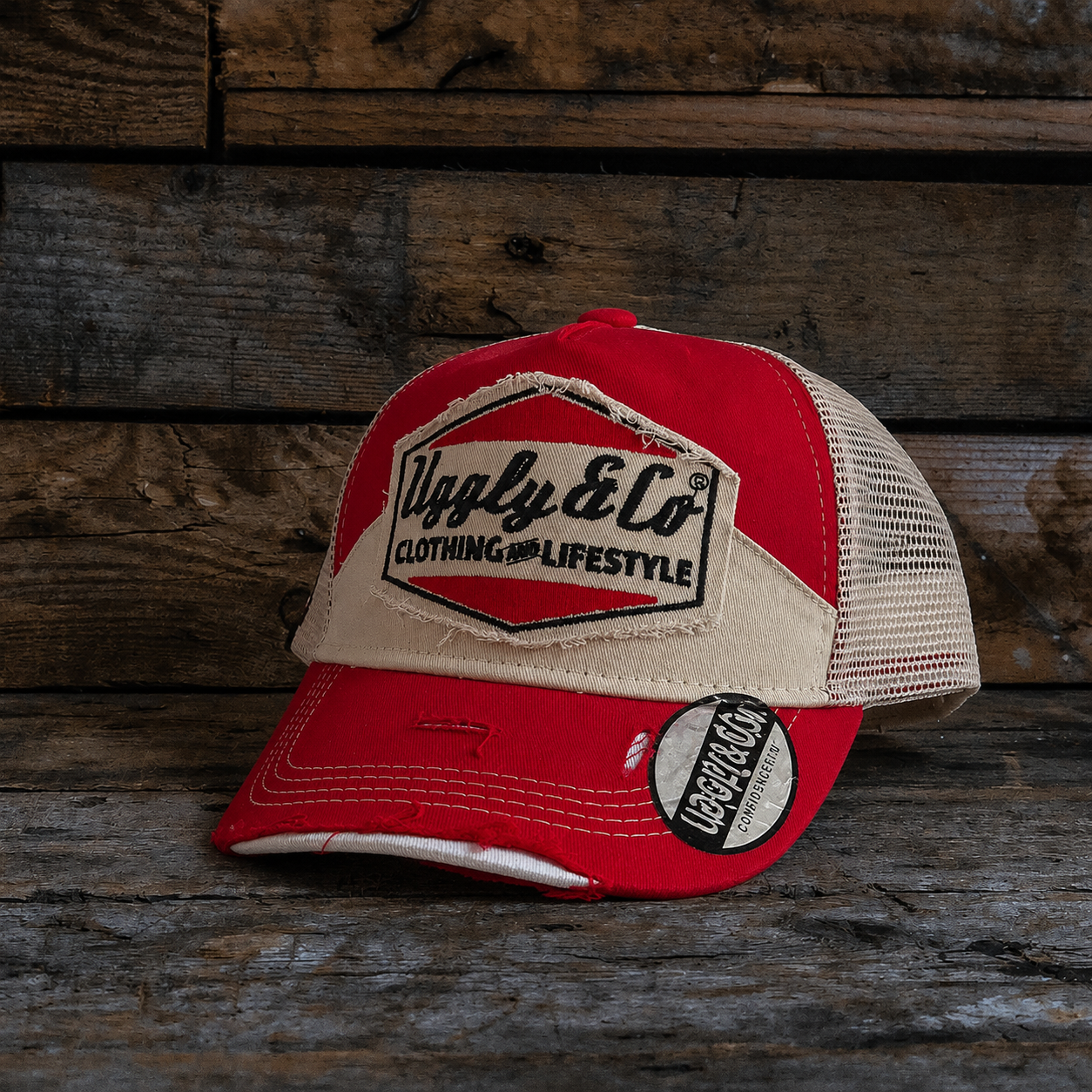 Red and beige trucker hat with Uggly&Co branding and a mesh back section on a wooden background