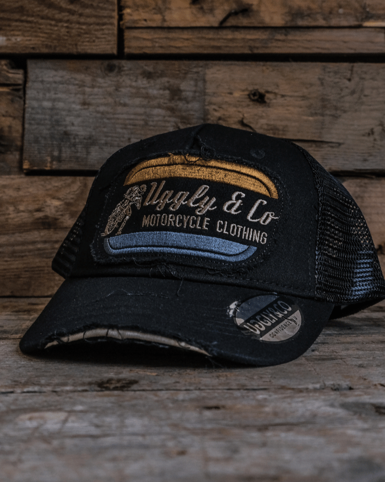 An all-black trucker hat with a vintage-inspired flat track racing logo in yellow and blue on the front with a wooden background