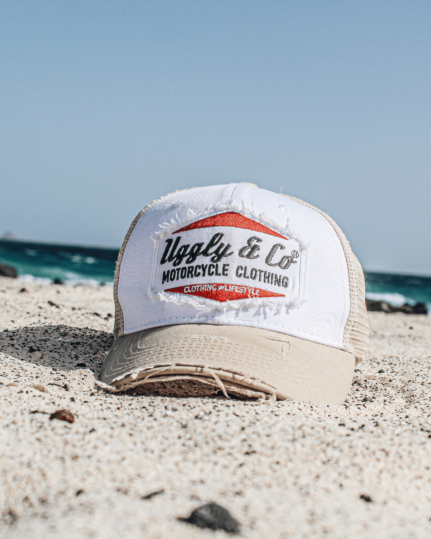 A beige and white Uggly & Co motorcycle clothing trucker hat with a mesh back and an embroidered logo on the front placed on the sand on a beach