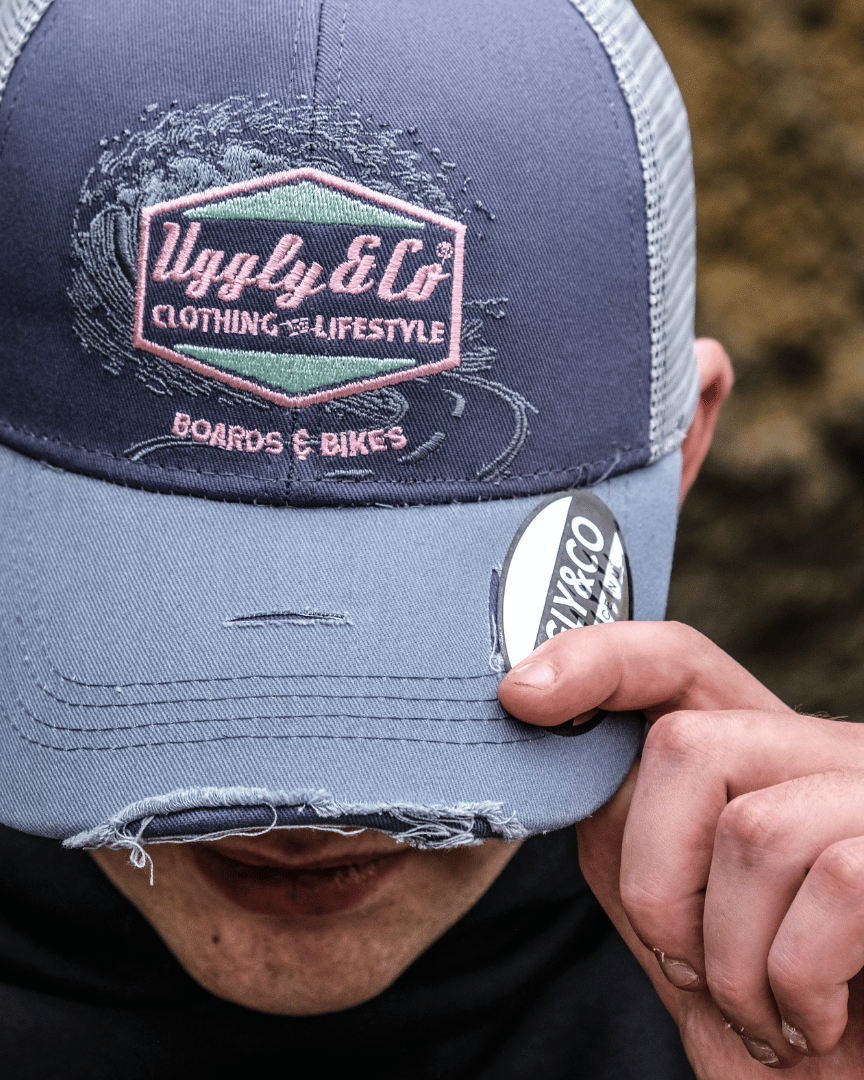 Front view of a model looking down while wearing a trucker hat with a purple front panel and a blue mesh back, featuring a pink diamond Uggly&Co logo and the text 'Boards & Bikes' with a blurred rocky background