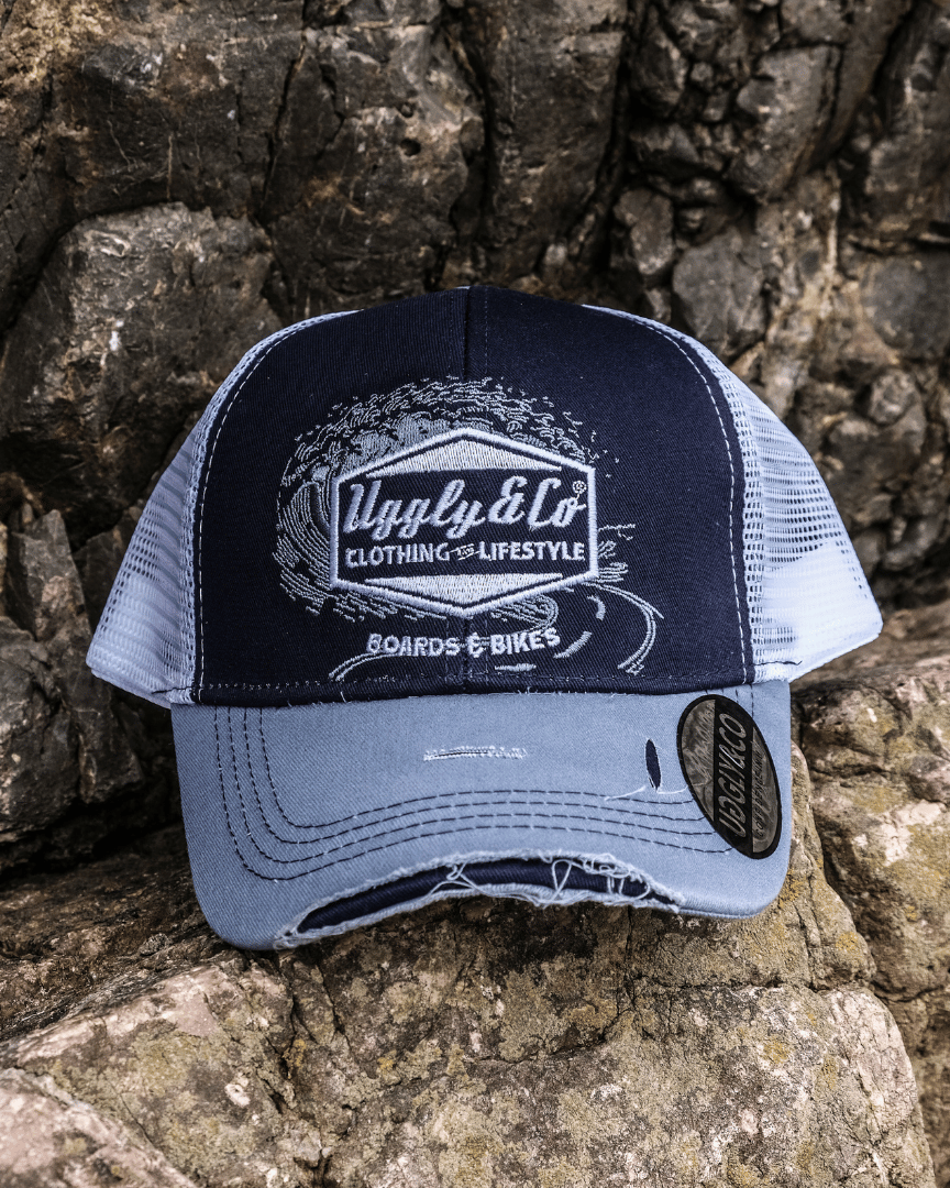 A blue trucker hat with a white print of the Uggly&Co diamond logo and a light blue wave texture on the front, featuring a navy blue front panel, a sun-faded light blue peak, and a breathable white mesh back, displayed on a rocky background