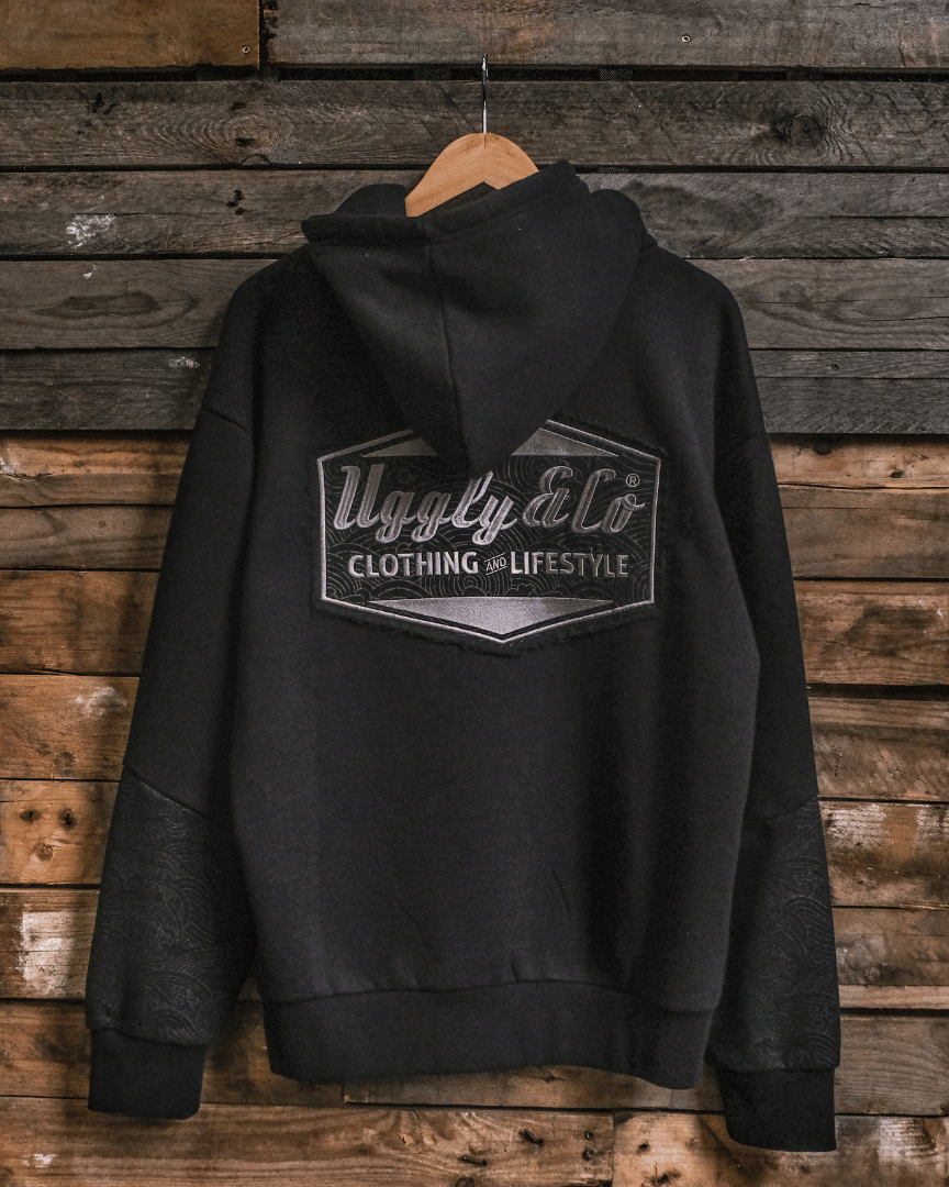 A black hoodie with a hood, featuring the Uggly&Co logo in grey embroidery on the front and a small woven label on the left sleeve. Displayed against a wooden backdrop.