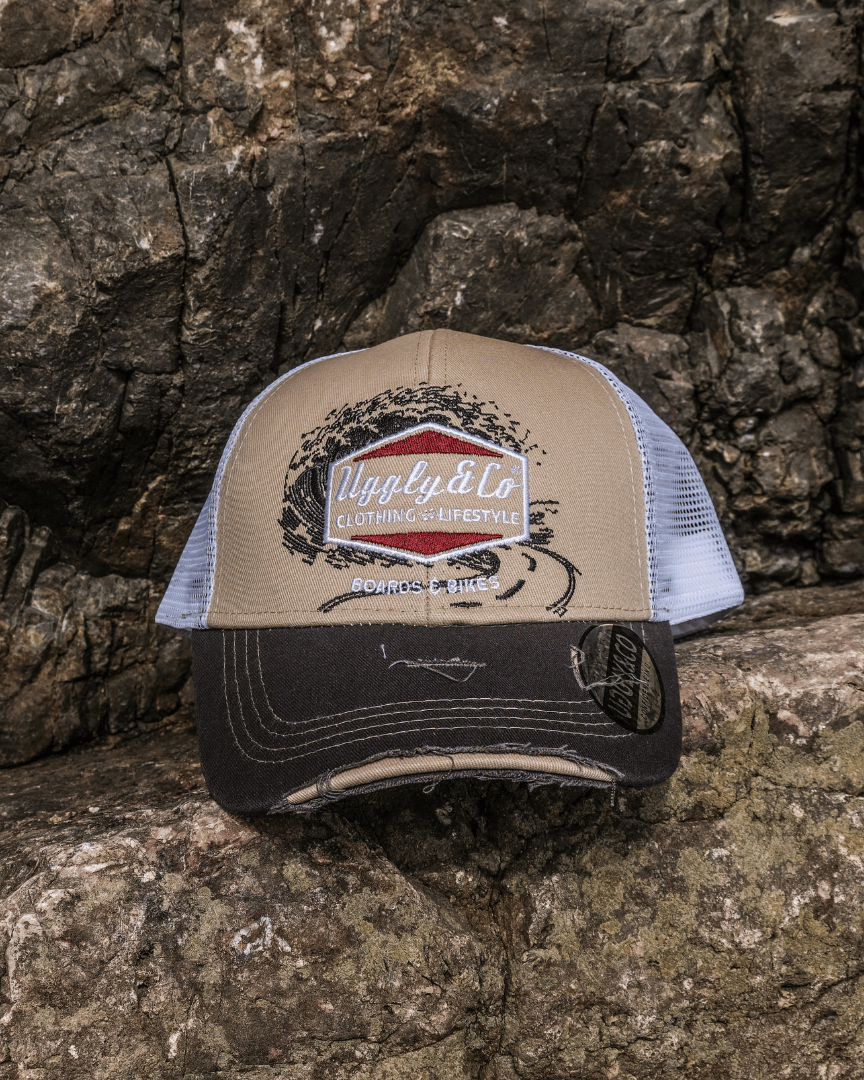 A beige and brown trucker cap with a diamond Uggly&Co logo and wave texture design, featuring a white mesh back and a curved peak, displayed on rocks
