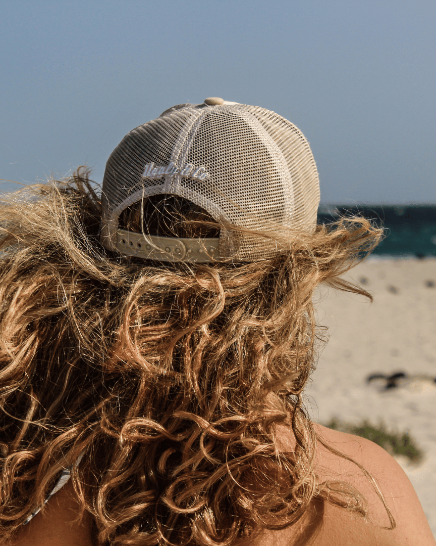 Rear view of a model with long hair on a beach wearing a beige and white Uggly & Co motorcycle clothing trucker hat with a mesh back and an embroidered logo on the front