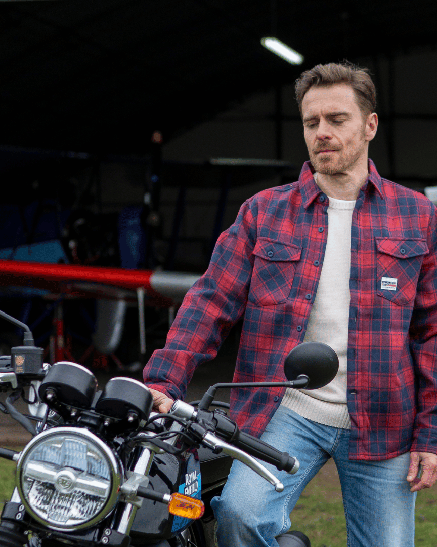 Man in a red plaid shirt standing next to a motorcycle with an airplane in the background.