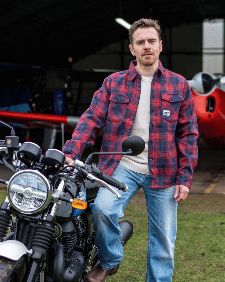 Man in a red plaid shirt standing next to a motorcycle with an airplane in the background.