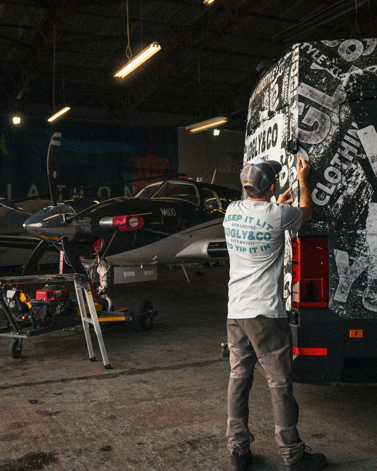 Uggly & Co Grey & White Vintage Diamond Trucker Cap worn by a man working on a van wrap in a hangar.