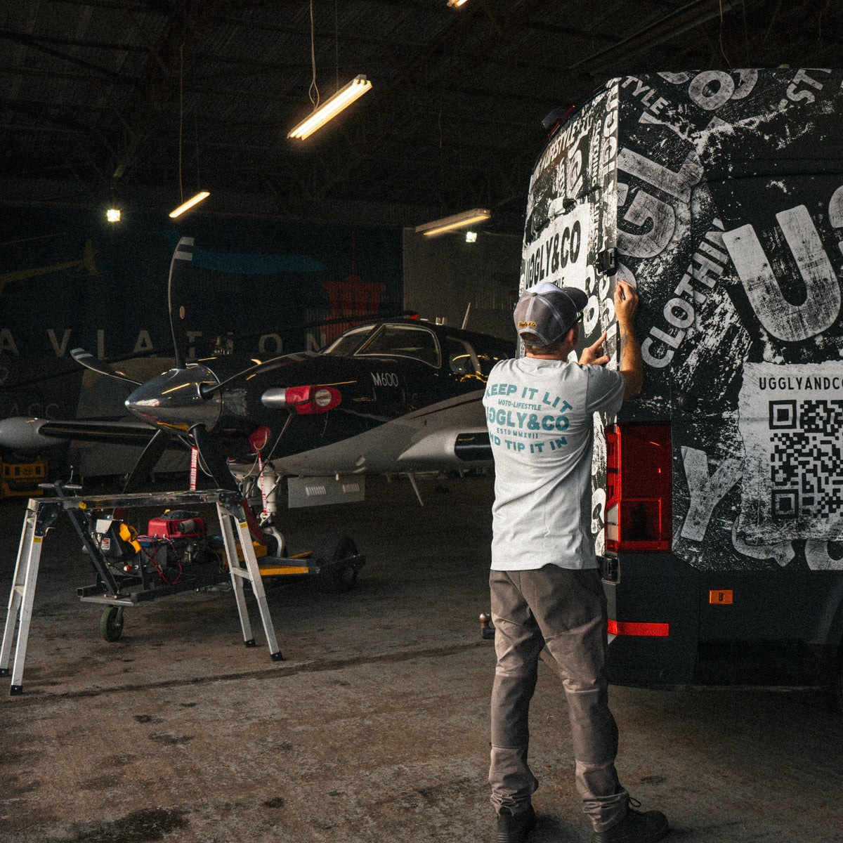 Uggly & Co Grey & White Vintage Diamond Trucker Cap worn by a man working on a van wrap in a hangar.
