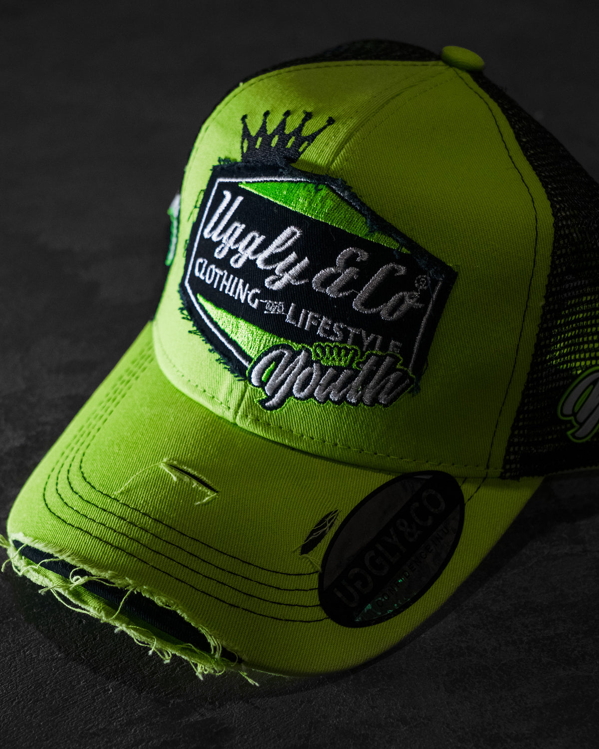 Angled product photo of the Uggly & Co Fluro Diamond Trucker Hat x Kyle Ryde on a concrete floor, highlighting the motorcycle lifestyle cap design.