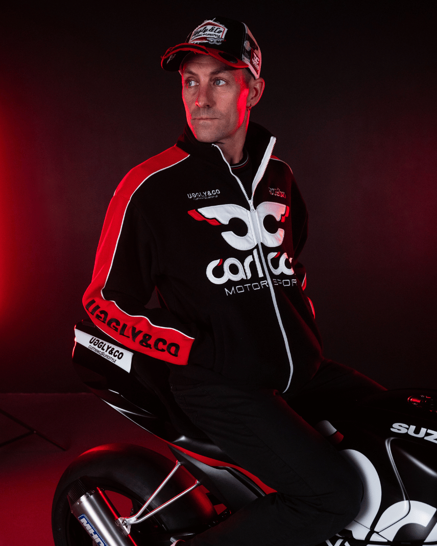 Josh Brookes sitting on his motorbike while wearing a black and red jacket and trucker hat with Carl Cox Motor Sport, Uggly&Co and Team Classic Suzuki branding