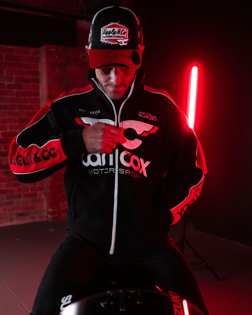 Front view of Josh Brookes sitting on his motorbike while wearing a black and red jacket and trucker hat with Carl Cox Motor Sport, Uggly&Co and Team Classic Suzuki branding