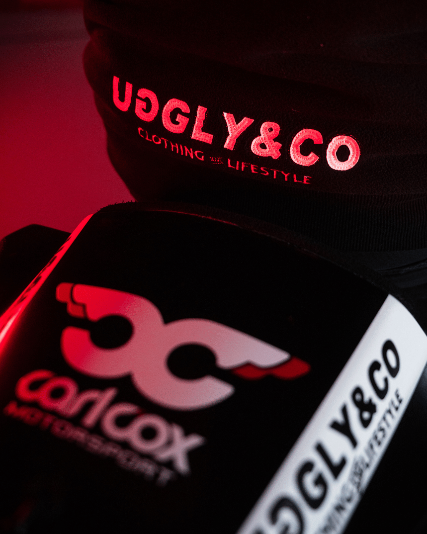 Close-up of a black jacket with 'Uggly&Co' and Carl Cox motorsport logos on a dark background