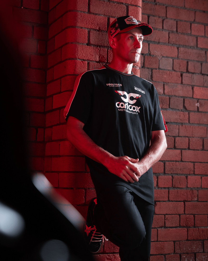 Josh Brookes wearing a Carl Cox motorsport and Team Classic Suzuki black and red t-shirt while leaning against a wall