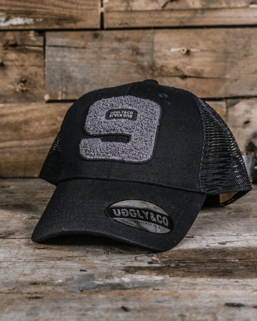 A black trucker cap with the number 9 and the text 'The Black Dub' on the front, laid on a wooden surface.