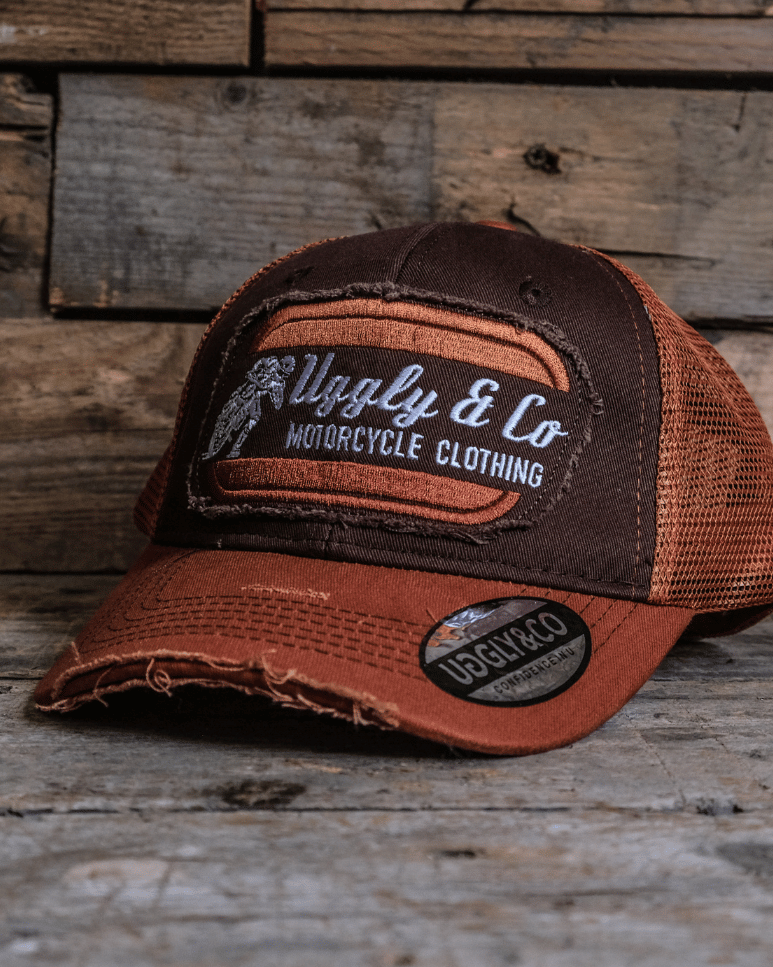 A brown trucker hat with an orange brim, featuring a flat track Uggly&Co logo patch on the front, placed on a wooden surface.