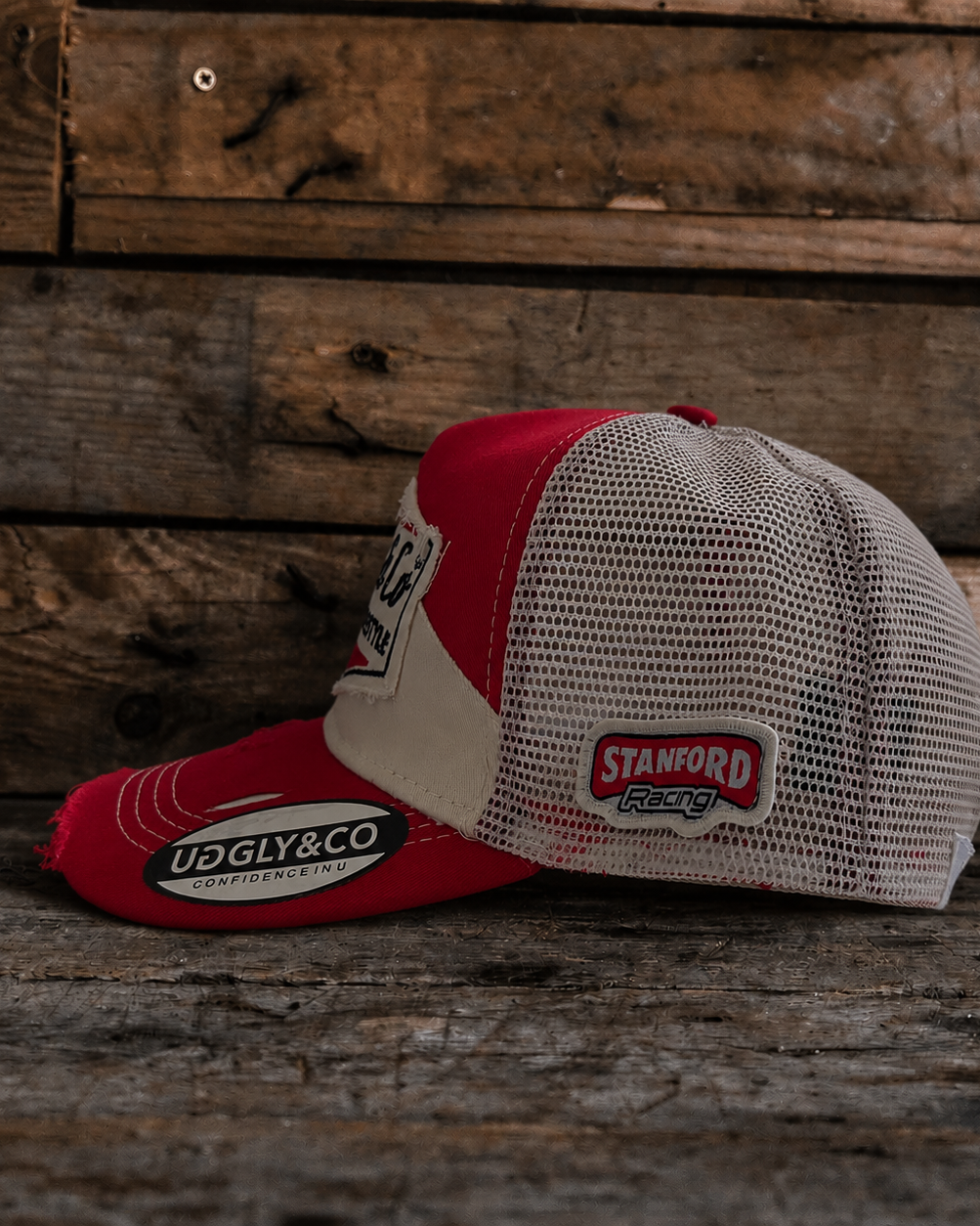 Side view of a red and beige trucker hat with Uggly&Co branding and a mesh back section on a wooden background