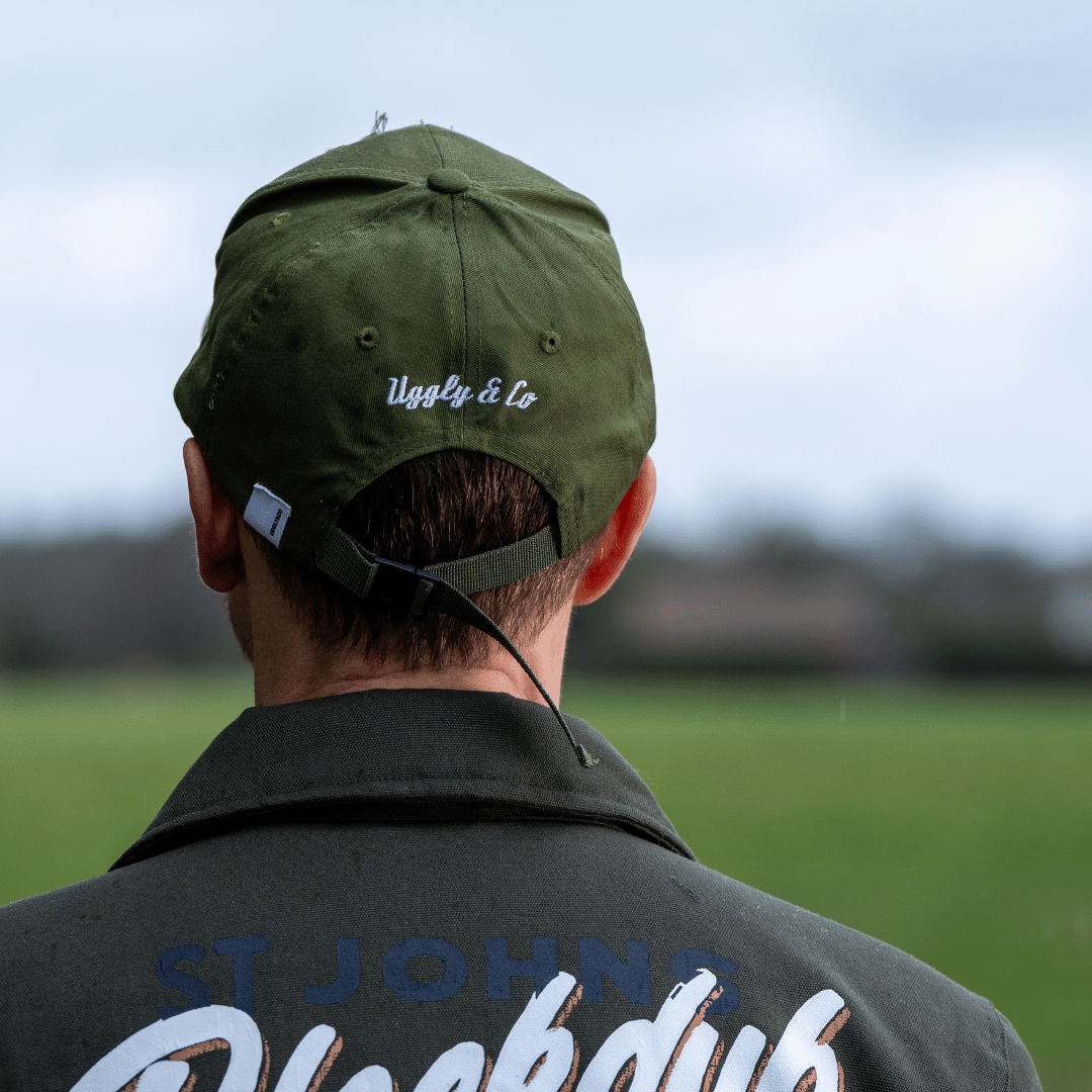 Rear view of a model wearing a green vintage cap with an Uggly&Co brand logo on the back and a blurred background
