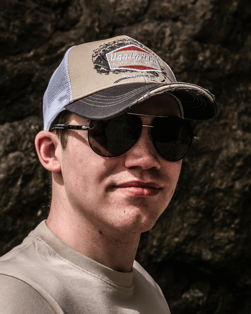 A model wearing sunglasses and a beige and brown trucker cap with a diamond Uggly&Co logo and wave texture design, featuring a white mesh back and a curved peak, with a blurred rocky background.