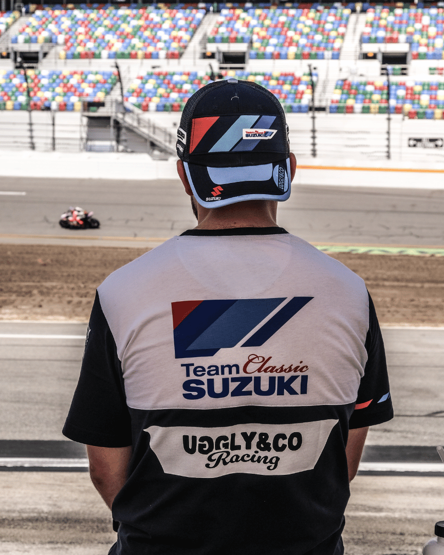Rear view of a model wearing a trucker hat backwards with geometric pattern featuring blue, red, and black colours, with the Team Classic Suzuki and Suzuki logos on it by a race track